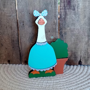 May include: A wooden goose figurine wearing a turquoise dress with white polka dots and a blue polka dot bow. The goose is standing on a green base and holding a potted green plant.