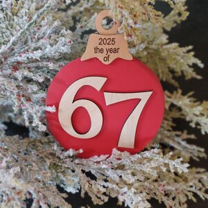 Könnte beinhalten: Ein roter Weihnachtsschmuck mit der Zahl 67 in Holzschrift. Der Schmuck hat eine Oberseite aus Holz mit dem Text "2025 the year of". Der Schmuck wird auf einem gefrosteten Tannenzweig präsentiert.