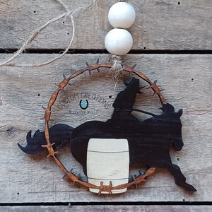 Op de afbeelding: Houten uitsnijding van een paard en ruiter die barrel racing doen, met een prikkeldraadkrans eromheen. Het paard en de ruiter zijn zwart, en de ton is wit. De krans is bruin.