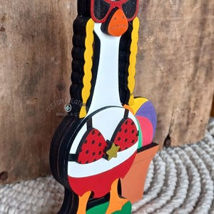 May include: A wooden goose figurine wearing red sunglasses, a red and white polka dot bikini, and a red skirt. The goose is standing on a green base with a yellow flower. The goose is holding a purple and white beach ball.