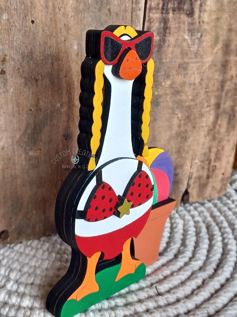May include: A wooden goose figurine wearing red sunglasses, a red and white polka dot bikini, and a red skirt. The goose is standing on a green base with a yellow flower. The goose is holding a purple and white beach ball.