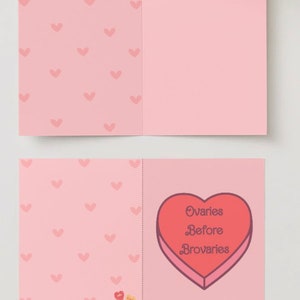 May include: A pink card with a heart-shaped box on the front. The box is red and has the text "Ovaries Before Brovaries" inside. The back of the card has a pattern of pink hearts.