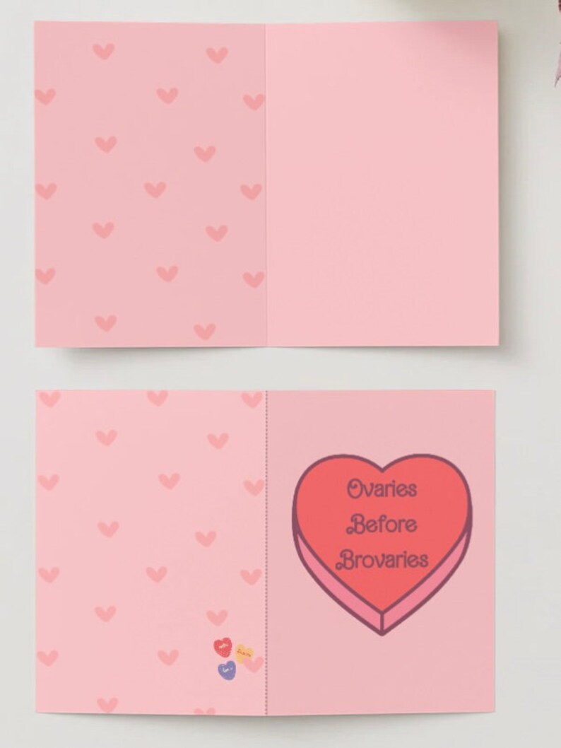 May include: A pink card with a heart-shaped box on the front. The box is red and has the text "Ovaries Before Brovaries" inside. The back of the card has a pattern of pink hearts.