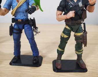 May include: Two action figures standing on black display bases. The figure on the left is wearing a blue sailor uniform, a rope belt, and holding a green parrot. The figure on the right is wearing a green and yellow camouflage uniform, a black shirt, and a brown hat. Both figures are holding weapons.