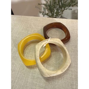 May include: Three square-shaped bangle bracelets. One is a solid dark brown, one is a solid bright yellow, and one is a cream-colored bangle with a subtle speckled pattern. Each bracelet features a smooth, polished finish and rounded corners.