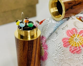Puede incluir: Un alfiletero de madera con tapa de latón y un patrón floral. El alfiletero está abierto y contiene una variedad de alfileres de cabeza de vidrio de colores.