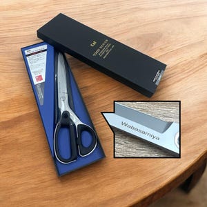 Może przedstawiać: Para nożyczek ze stali nierdzewnej Kai serii 7000 z czarnymi uchwytami w niebiesko-czarnym pudełku. Nożyczki są grawerowane nazwą "Wabasamiya".