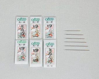 Op de afbeelding: Zes pakjes Clover naalden, elk pakje met het label "ぬい針" (uitgesproken als "nui bari"), wat "naald" betekent in het Japans. De pakjes zijn wit met groene en rode accenten. Er zijn ook zeven gouden naalden op een witte achtergrond.