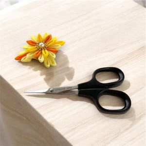 May include: Black handled scissors with a silver blade. The scissors are labeled "CANARY JAPAN" and "STAINLESS STEEL". A yellow and orange flower with a pearl in the center is in the background.