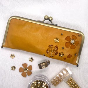 May include: A mustard yellow leather wallet with a brass clasp and floral embellishments. The wallet is decorated with gold-toned sequins and rhinestones.