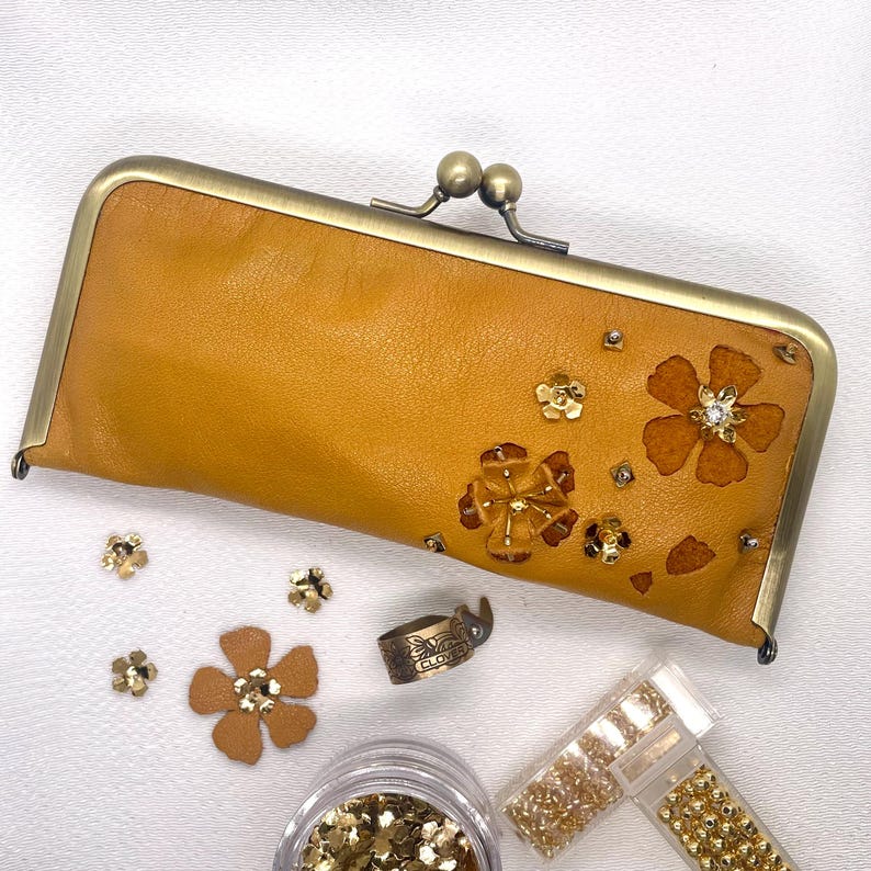 May include: A mustard yellow leather wallet with a brass clasp and floral embellishments. The wallet is decorated with gold-toned sequins and rhinestones.