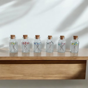 May include: Seven small glass jars with cork tops filled with colorful glass headed pins. The jars are lined up on a wooden shelf.