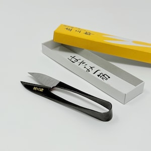 Peut inclure: Ciseaux japonais noirs et argentés pour fleurs avec une boîte jaune et blanche. Les ciseaux ont une poignée incurvée et des lames tranchantes. La boîte contient du texte japonais.