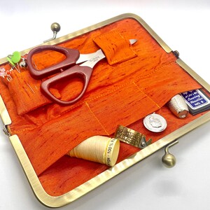 May include: A gold metal sewing kit with a bright orange fabric interior. The kit contains a pair of small scissors, a thimble, a spool of thread, a needle case, and a few sewing pins.