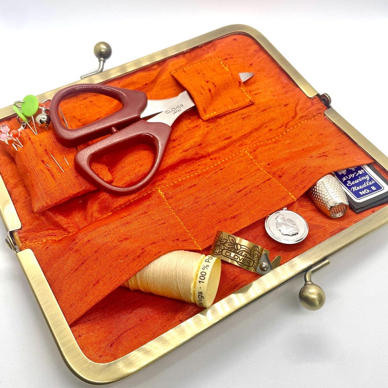 May include: A gold metal sewing kit with a bright orange fabric interior. The kit contains a pair of small scissors, a thimble, a spool of thread, a needle case, and a few sewing pins.