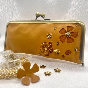 May include: A mustard yellow leather clutch purse with a brass clasp and floral cutouts decorated with gold and clear rhinestones.