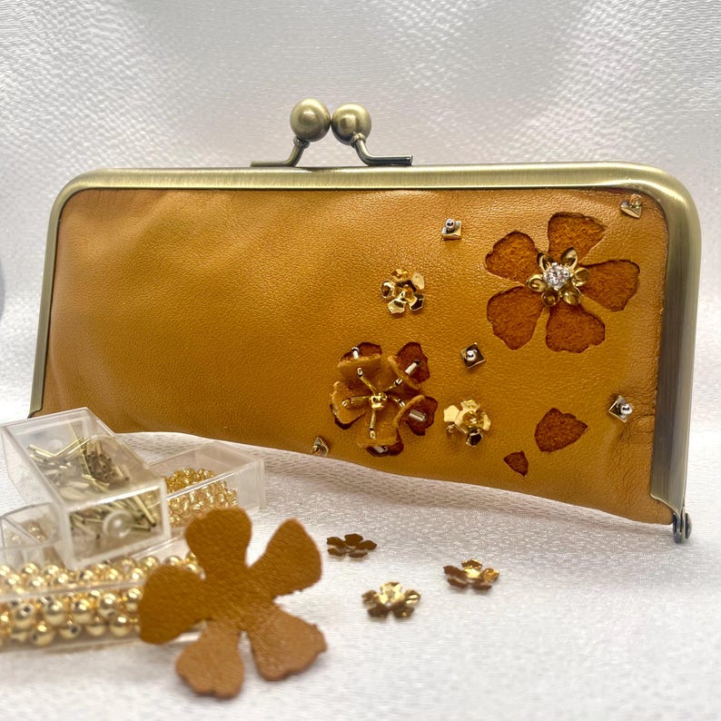 May include: A mustard yellow leather clutch purse with a brass clasp and floral cutouts decorated with gold and clear rhinestones.