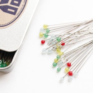 May include: A collection of colorful sewing pins with round, beaded heads in red, yellow, green, and clear. The pins are scattered on a white surface next to an open, silver metal tin. The tin has text on the lid.