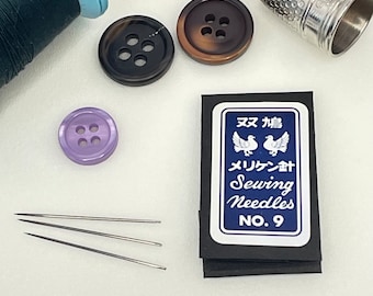 Op de afbeelding: Een klein doosje met naalden voor het naaien met de tekst "Esu Kaeru" en "Meriken Goshi Sewing Needles No. 9". De doos is blauw met witte tekst en twee witte duiven. Op de afbeelding zijn vier naalden voor het naaien en drie knopen te zien.