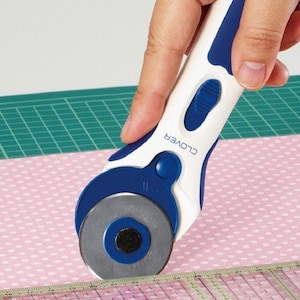 Puede incluir: Un cortador rotatorio blanco y azul con la palabra "CLOVER" impresa en el mango, cortando tela rosa con lunares. Una regla transparente es visible en la parte inferior. Herramienta de costura.