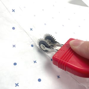 May include: A red handled tracing wheel with two spiked wheels. The tool is used for transferring pattern markings onto fabric. The background is white with blue cross and dot patterns.