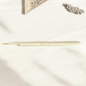 Peut inclure: Un outil blanc et fin avec une pointe dorée. L'outil est étiqueté "Magical Pick".