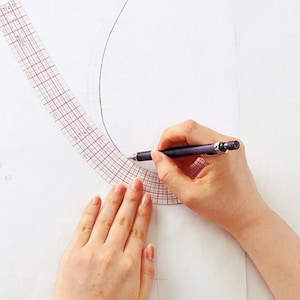 Puede incluir: Una regla de plástico transparente con un patrón de cuadrícula se utiliza para dibujar una línea curva en una superficie blanca. Las manos de una persona sostienen un lápiz y la regla.