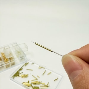 May include: A close-up of a small, gold-colored needle held in a hand. The needle is thin and has a sharp point. Several small, gold-colored metal pieces are in clear plastic containers in the background. The items appear to be craft supplies.