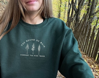 Pode incluir: Uma camisa de frio verde escura com o texto "The Sound of Rain Amongst the Pine Trees" e um gráfico de três pinheiros.