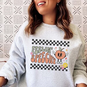May include: Gray sweatshirt with a peach graphic and the text "Feelin' Speechy" in a retro font. The peach has a face and is holding up peace signs. The text is surrounded by a black and white checkered border and flowers.