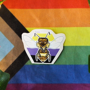 Könnte beinhalten: Ein Aufkleber mit einer Bienenillustration auf weißem Hintergrund. Die Biene ist gelb und schwarz mit lila und schwarzen Streifen dahinter. Der Text "Enbee" befindet sich oben auf dem Aufkleber. Der Aufkleber befindet sich auf einer Regenbogen-Pride-Flagge.