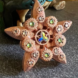 May include: A copper-colored star-shaped ornament with a detailed design and iridescent green and clear rhinestones.