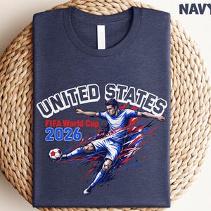 May include: Heather navy t-shirt featuring a soccer player kicking a ball, with the text "UNITED STATES FIFA World Cup 2026". The design incorporates red, white, and blue colors, and the shirt is folded on a woven mat.