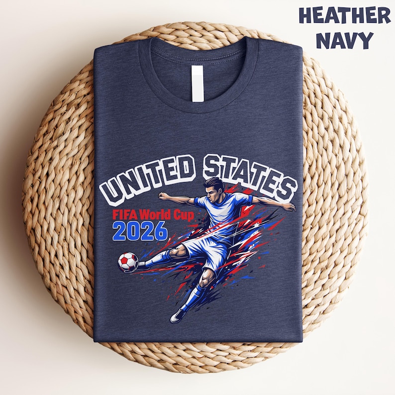 May include: Heather navy t-shirt featuring a soccer player kicking a ball, with the text "UNITED STATES FIFA World Cup 2026". The design incorporates red, white, and blue colors, and the shirt is folded on a woven mat.