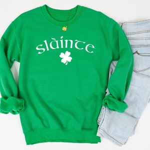 Op de afbeelding: Groen sweatshirt met witte tekst die "sl&aacute;inte" zegt en een wit vierbladig klavertje.