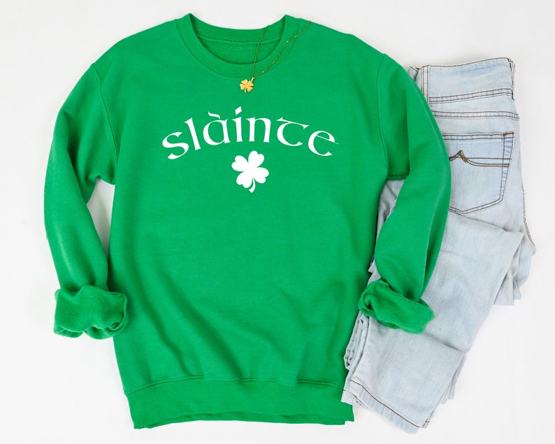 Op de afbeelding: Groen sweatshirt met witte tekst die "sl&aacute;inte" zegt en een wit vierbladig klavertje.