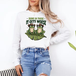 Mint green graphic t-shirt featuring two grumpy frogs in cowboy hats on a leaf with the sarcastic quote “Hang in there, it gets worse,” perfect for lovers of dark humor and quirky style.