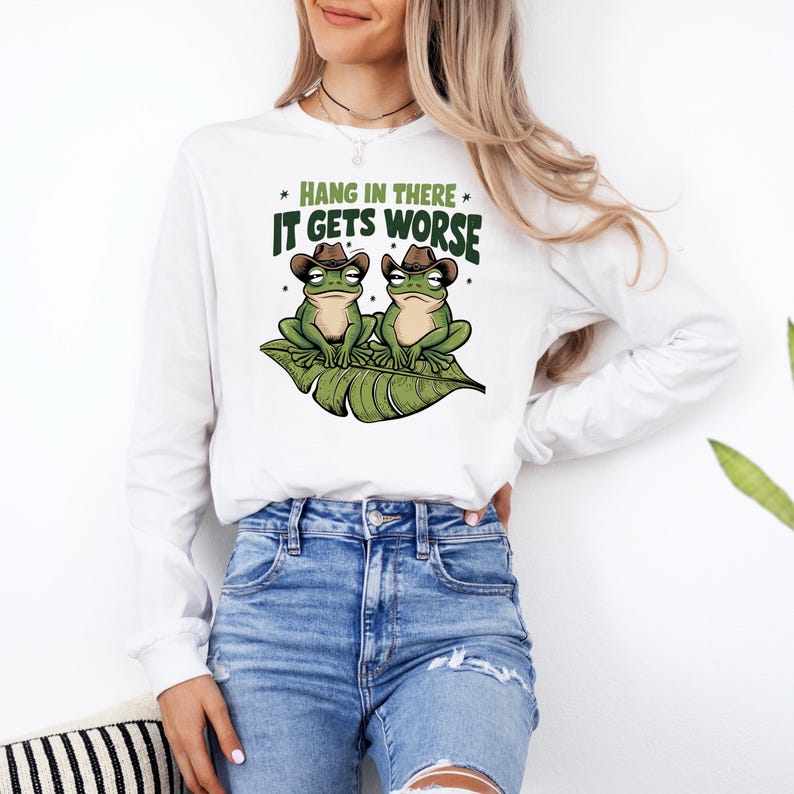 Mint green graphic t-shirt featuring two grumpy frogs in cowboy hats on a leaf with the sarcastic quote “Hang in there, it gets worse,” perfect for lovers of dark humor and quirky style.