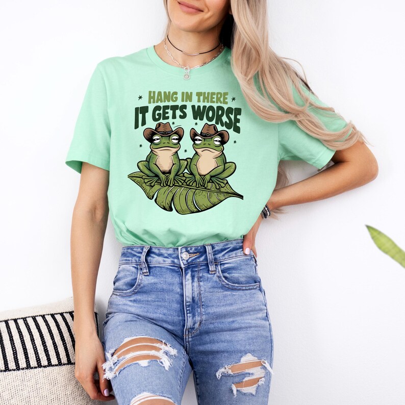 Mint green graphic t-shirt featuring two grumpy frogs in cowboy hats on a leaf with the sarcastic quote “Hang in there, it gets worse,” perfect for lovers of dark humor and quirky style.
