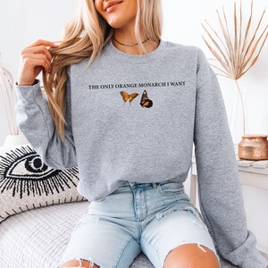 May include: A light gray sweatshirt with the text "THE ONLY ORANGE MONARCH I WANT" above two butterfly illustrations. One butterfly is orange, and the other is black and orange. The sweatshirt is a casual style.