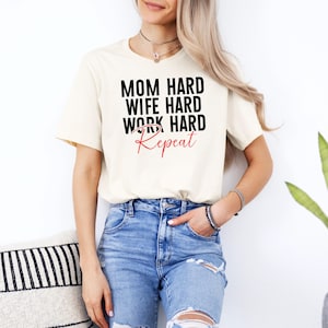 May include: Cream-colored t-shirt with the text "MOM HARD WIFE HARD WORK HARD Repeat" in black and red lettering. The model is wearing blue jeans. The shirt is a casual, everyday wear item.