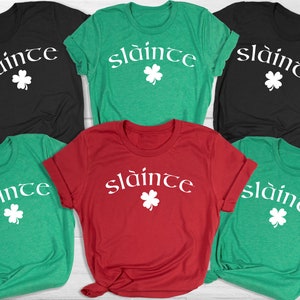 Op de afbeelding: Vijf T-shirts in verschillende kleuren met de Ierse zin "sl&aacute;inte" en een klavertje vier bedrukt op de voorkant.