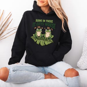 Mint green graphic t-shirt featuring two grumpy frogs in cowboy hats on a leaf with the sarcastic quote “Hang in there, it gets worse,” perfect for lovers of dark humor and quirky style.
