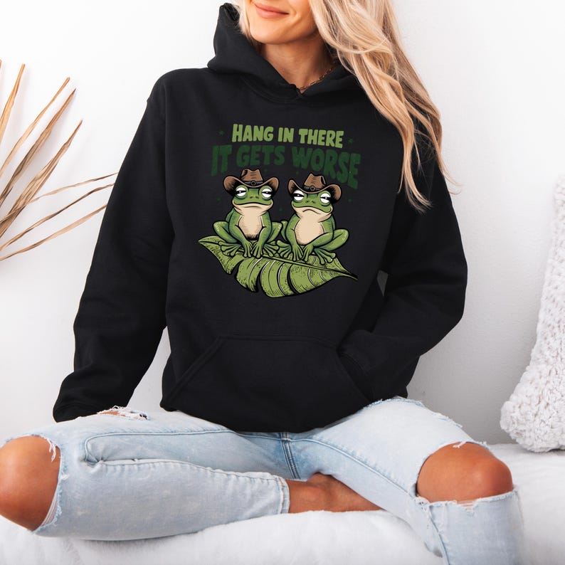 Mint green graphic t-shirt featuring two grumpy frogs in cowboy hats on a leaf with the sarcastic quote “Hang in there, it gets worse,” perfect for lovers of dark humor and quirky style.