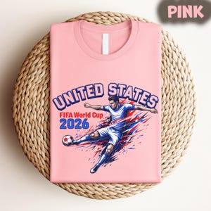 May include: A pink t-shirt featuring a graphic of a soccer player kicking a ball, with the text "UNITED STATES FIFA World Cup 2026" in blue and red. The shirt is folded on a woven, circular surface.