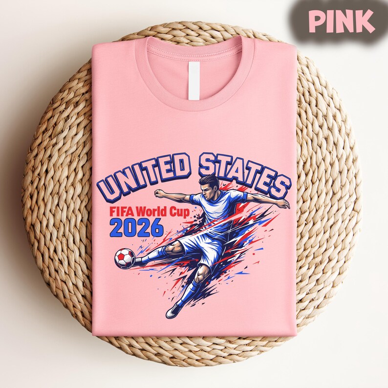 May include: A pink t-shirt featuring a graphic of a soccer player kicking a ball, with the text "UNITED STATES FIFA World Cup 2026" in blue and red. The shirt is folded on a woven, circular surface.