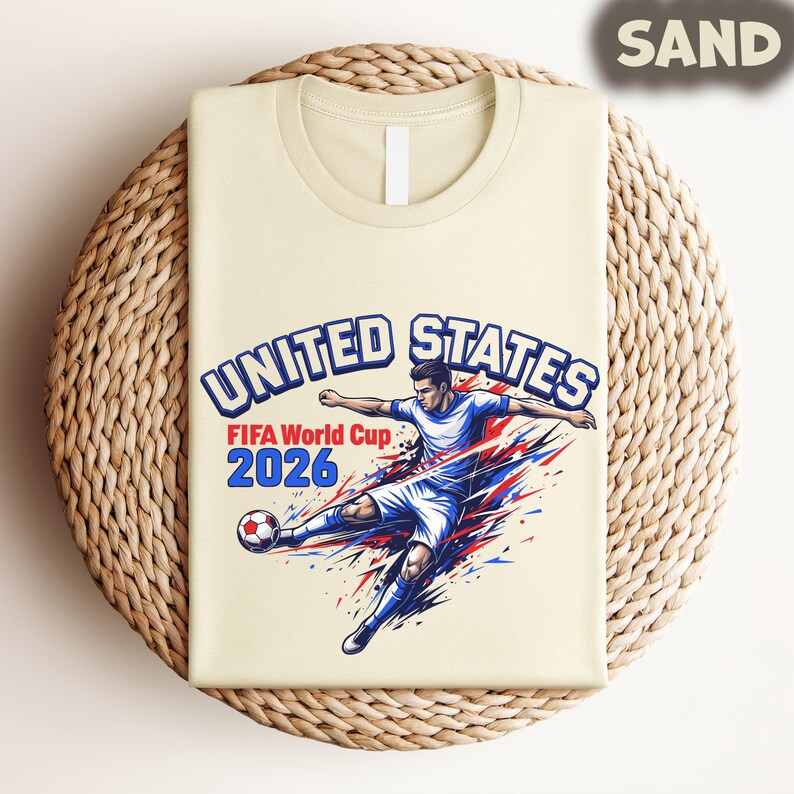 May include: Sand-colored t-shirt featuring a soccer player kicking a ball, with the text "UNITED STATES FIFA World Cup 2026" in blue and red. The design is a graphic illustration of a soccer player in action.