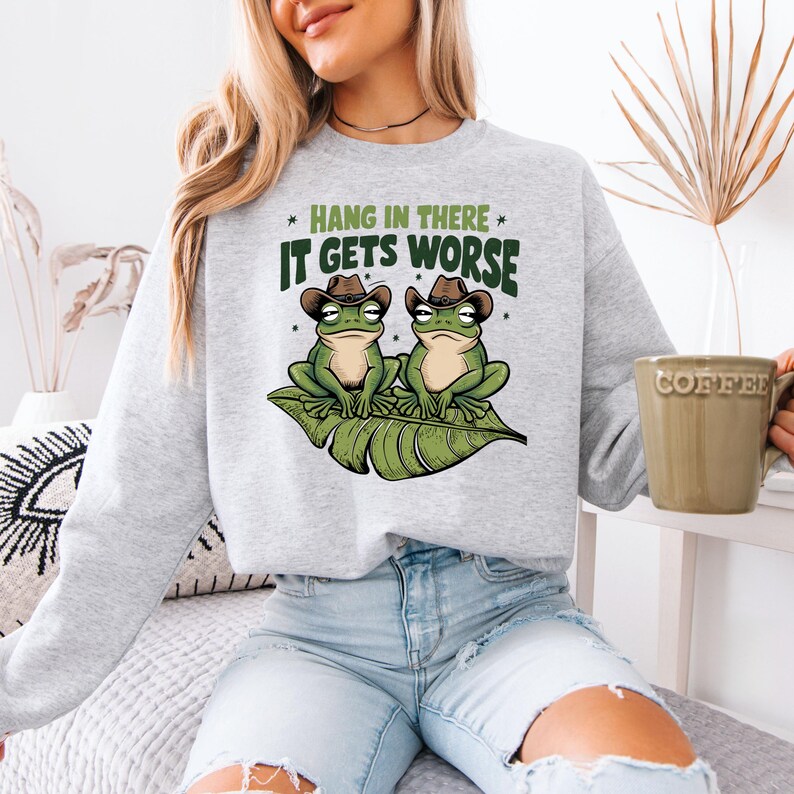 Mint green graphic t-shirt featuring two grumpy frogs in cowboy hats on a leaf with the sarcastic quote “Hang in there, it gets worse,” perfect for lovers of dark humor and quirky style.