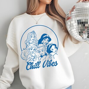 Minimalist blue line art of Disney princesses with "Chill Vibes" text on cream t-shirt; features Ariel, Cinderella, Snow White, Belle, and Jasmine in a relaxed, modern design.
