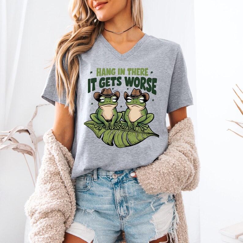 Mint green graphic t-shirt featuring two grumpy frogs in cowboy hats on a leaf with the sarcastic quote “Hang in there, it gets worse,” perfect for lovers of dark humor and quirky style.